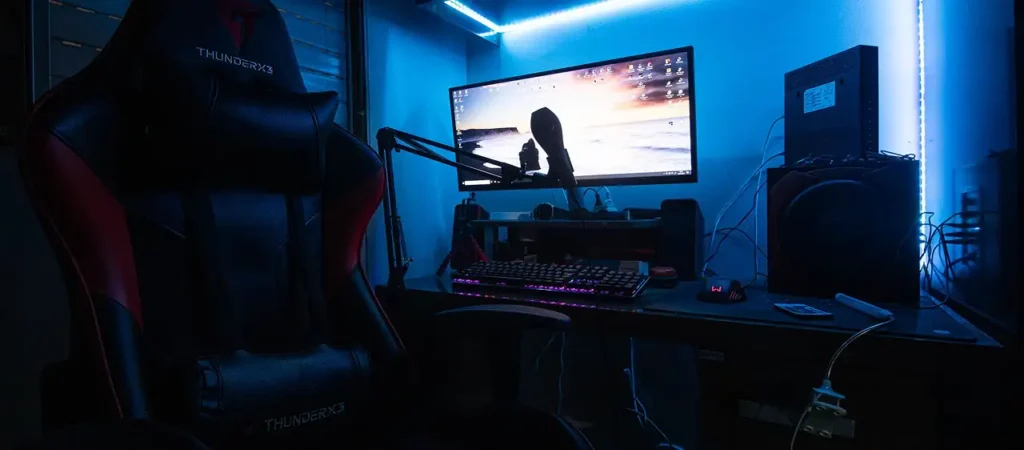 Configuração moderna de jogos RGB com uma cadeira de jogos, um monitor panorâmico, um teclado mecânico, um microfone e iluminação LED numa sala dedicada aos jogos.